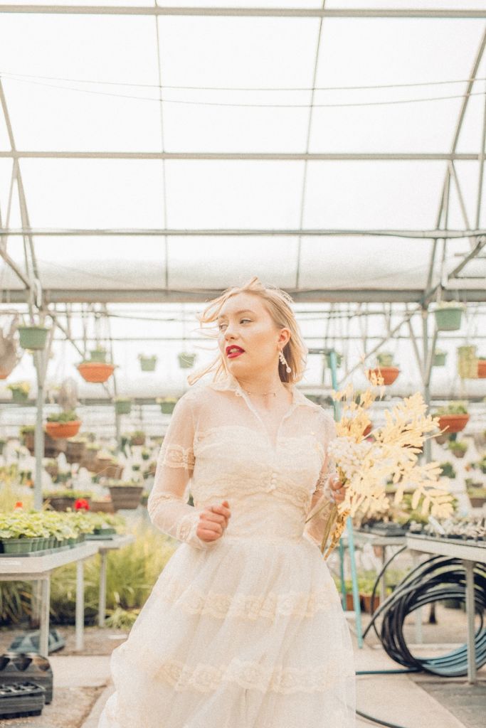 This vintage wedding dress is a unique, tea-length dress with a sheer jacket, lace stripes cover the jacket, arms, and the dress. The model runs looking to her side with yellow florals in her arms and in yellow shoes. Definitely ahead for its time with the transparent jacket. Model: Emily Johnson Dress: Wed Utah
